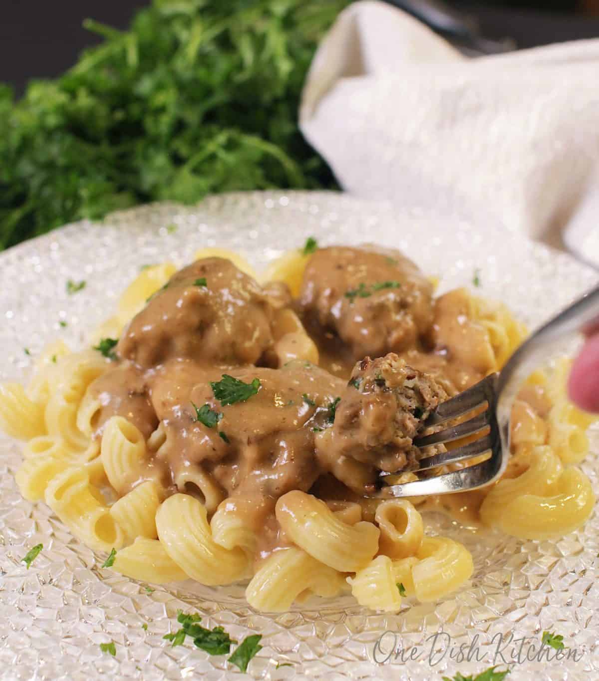 A fork picking up a meatball from a plate of buttered noodles topped with Swedish meatballs.