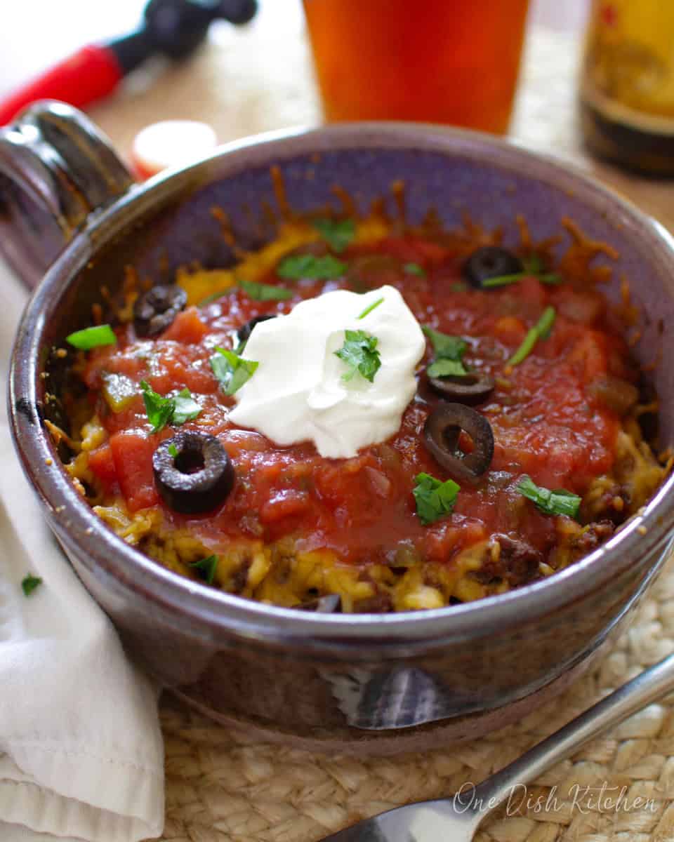 Taco casserole topped with salsa, sliced black olives, and a dollop of sour cream in a circular dish next to a pint of beer.