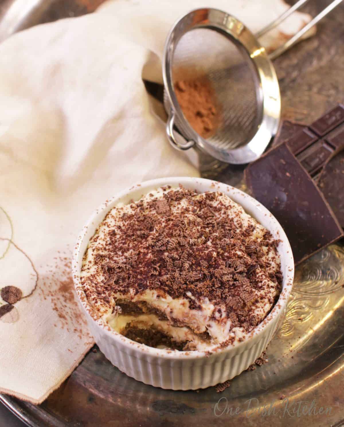 A mini tiramisu topped with chocolate shavings in a ramekin next to a piece of a chocolate bar, a cream-colored cloth napkin, and cocoa powder in a sieve all on a metal tray.