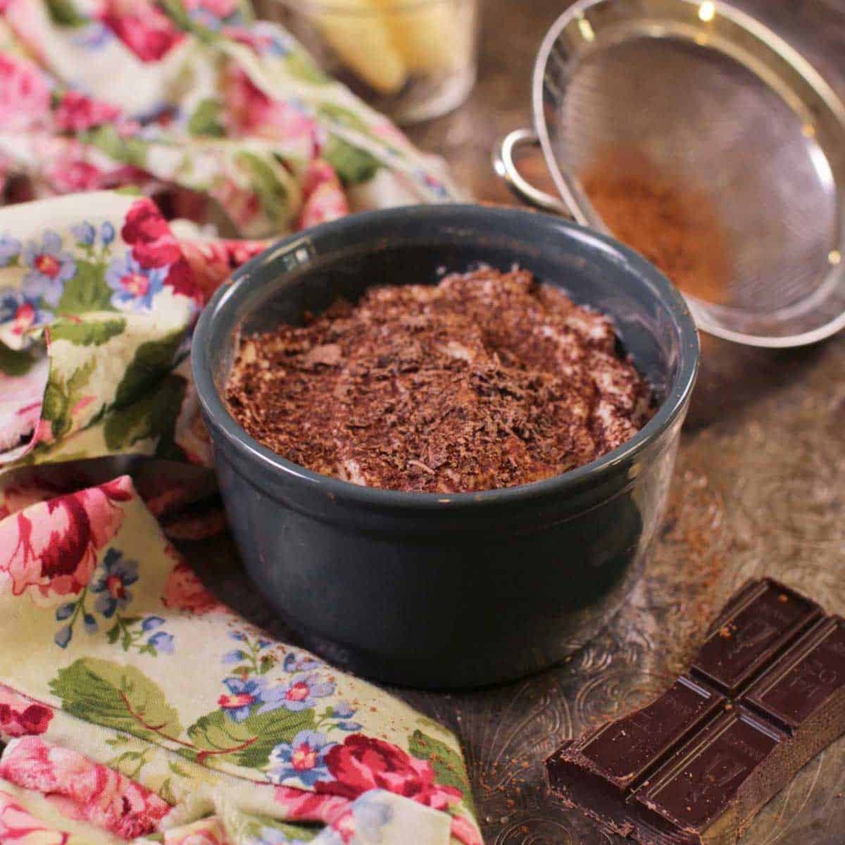 A mini tiramisu topped with chocolate shavings in a circular dish on a metal tray with a floral cloth napkin, a piece of a chocolate bar, and cocoa powder in a sieve.