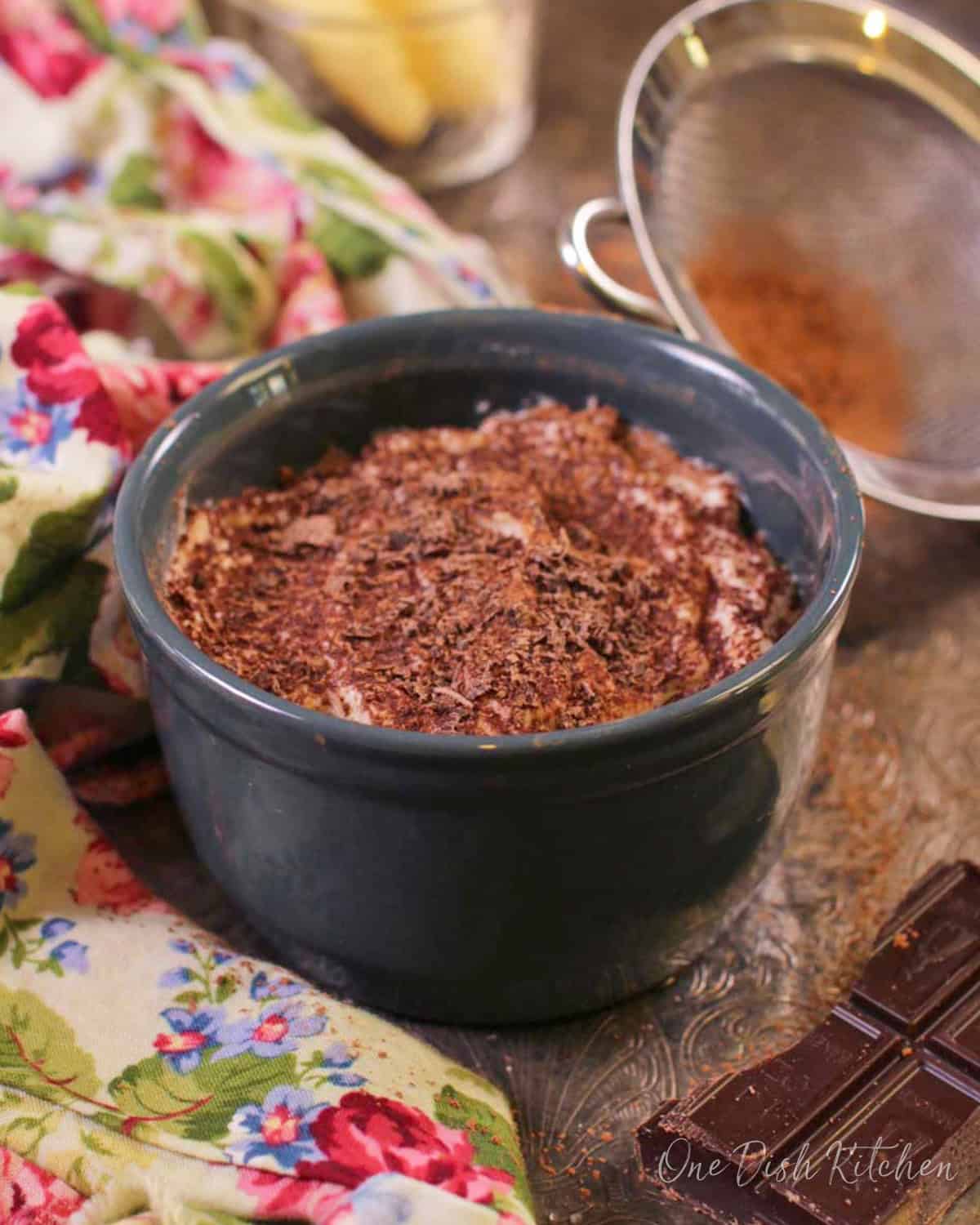 A mini tiramisu topped with chocolate shavings in a circular dish on a metal tray with a floral cloth napkin, a piece of a chocolate bar, and cocoa powder in a sieve.