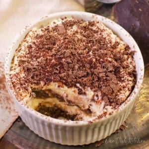 A mini tiramisu topped with chocolate shavings in a ramekin next to a piece of a chocolate bar, a cream-colored cloth napkin, and cocoa powder in a sieve all on a metal tray.