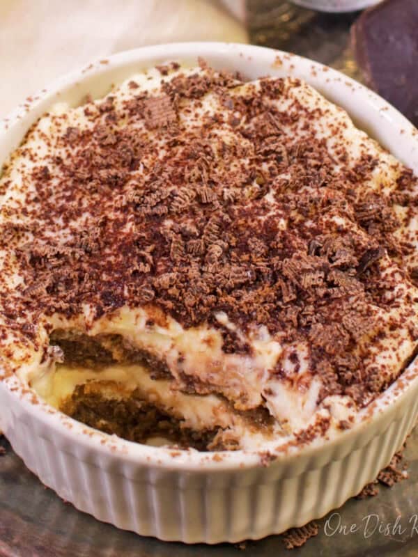 A mini tiramisu topped with chocolate shavings in a ramekin next to a piece of a chocolate bar, a cream-colored cloth napkin, and cocoa powder in a sieve all on a metal tray.