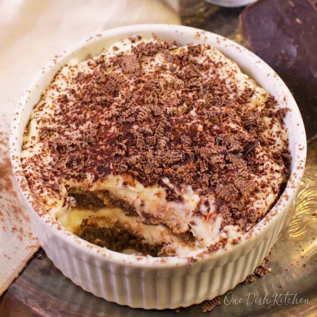 A mini tiramisu topped with chocolate shavings in a ramekin next to a piece of a chocolate bar, a cream-colored cloth napkin, and cocoa powder in a sieve all on a metal tray.