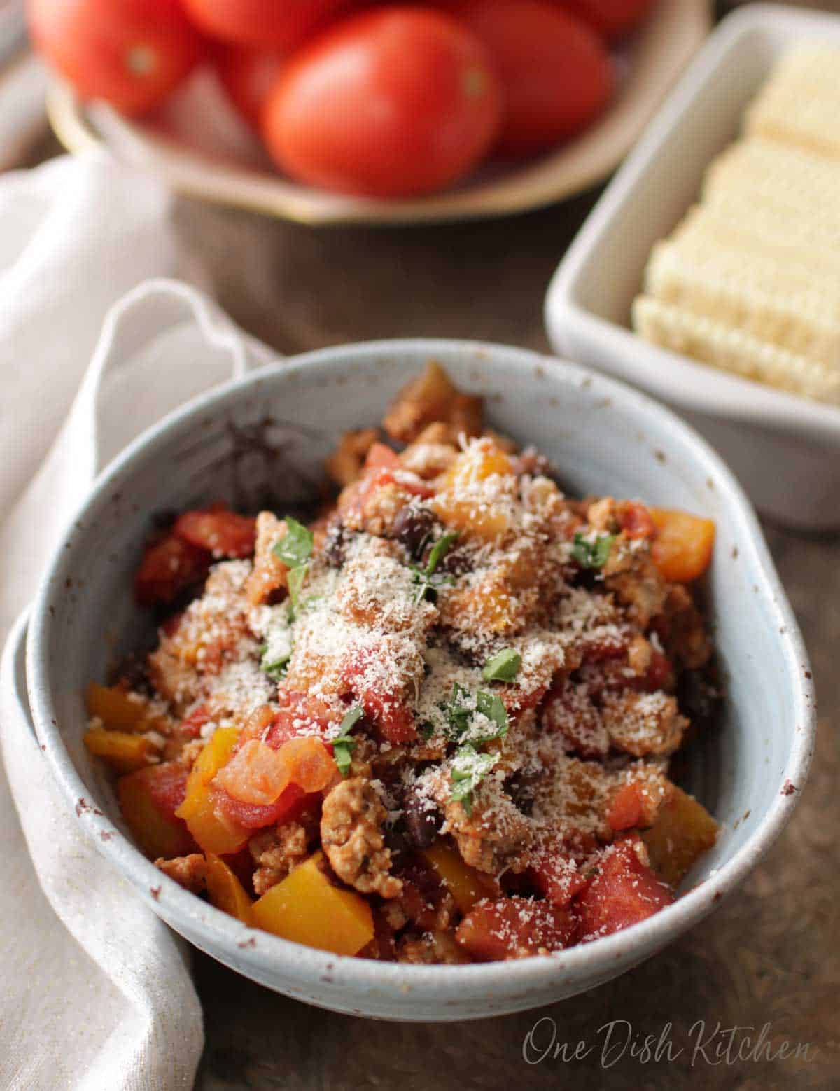 An overhead view of a bowl turkey chili topped with grated parmesan cheese in a blue bowl next to a plate of ripe red tomatoes. 