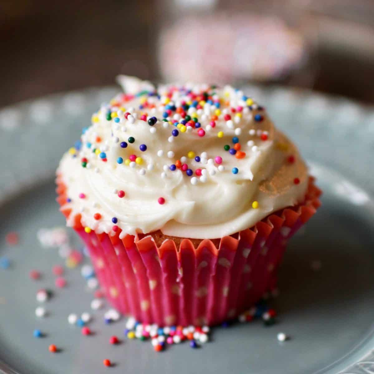 One cupcake topped with vanilla frosting and rainbow colored sprinkles on a plate.