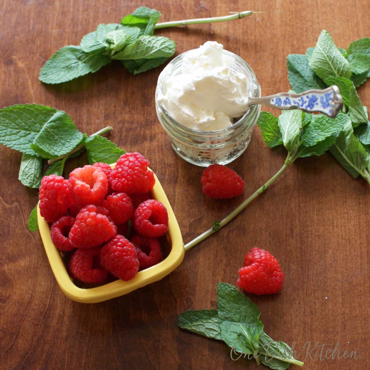 Raspberries in a bowl and homemade whipped cream in a small bowl with sprigs of fresh mint scattered on a table 