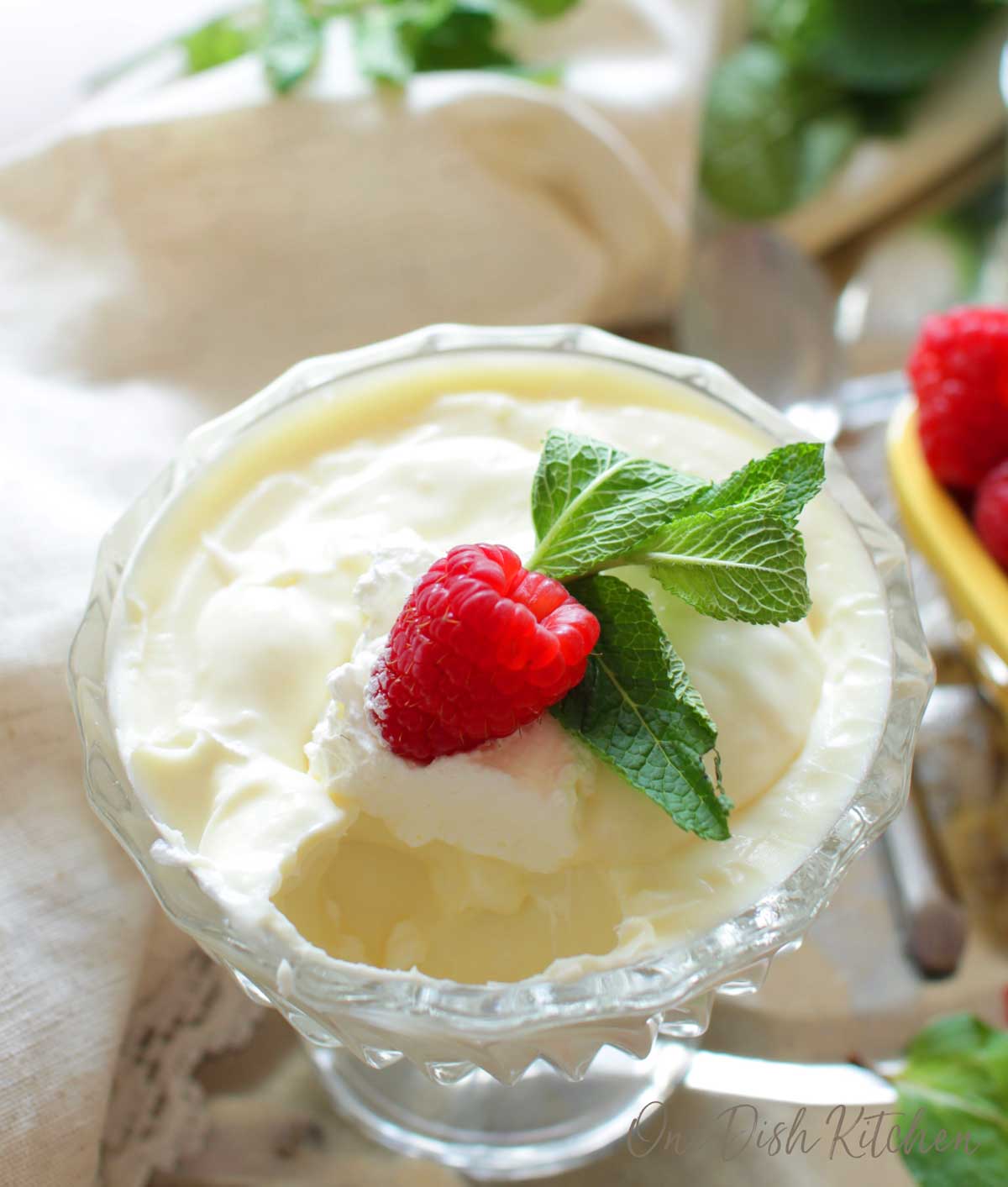 A half eaten bowl of vanilla pudding topped with whipped cream, a raspberry, and mint leaves in a dessert glass