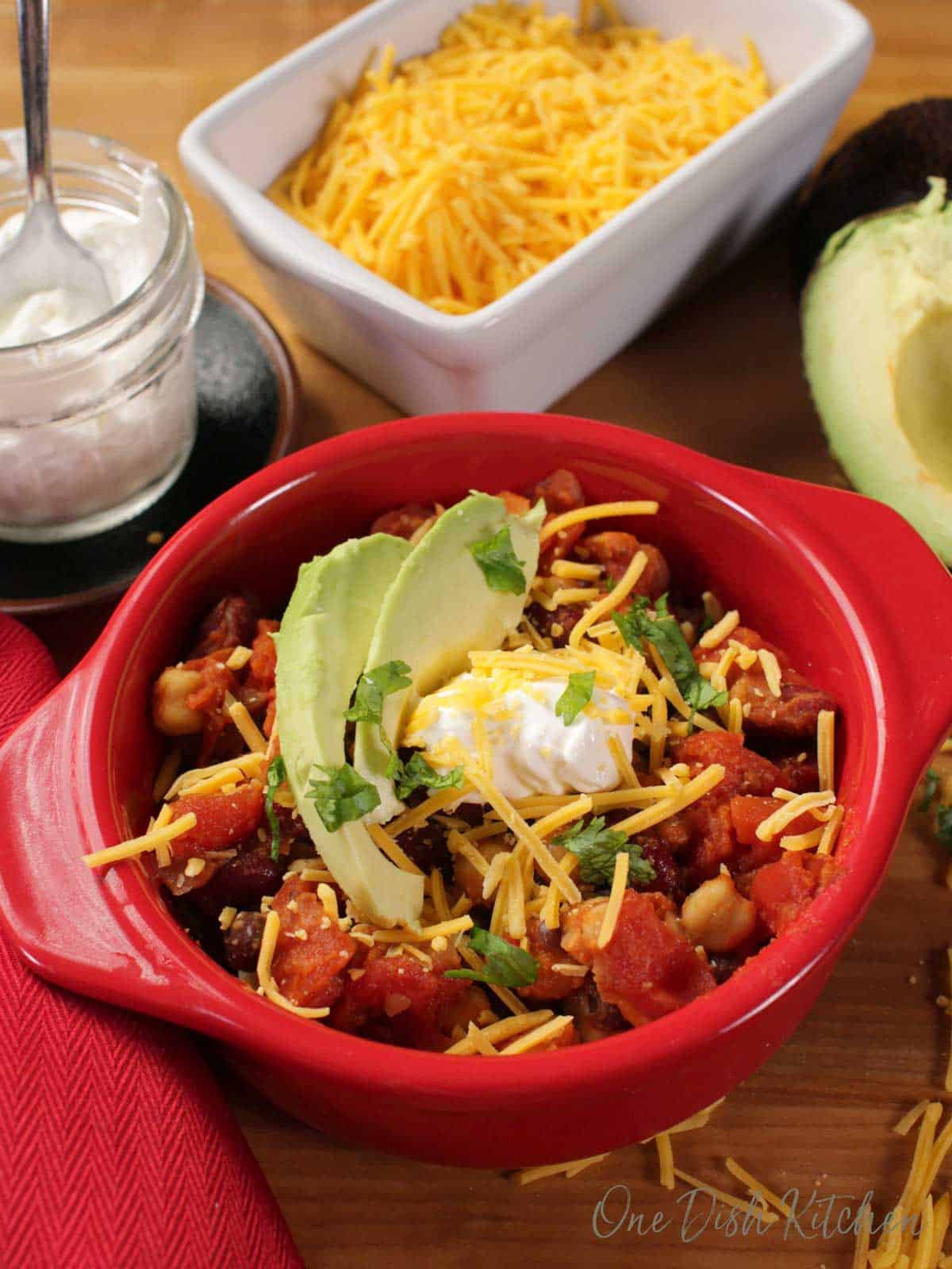 A red circular baking dish full of chili with beans, onions, and tomatoes, topped with two avocado slices, shredded cheese, and a dollop of sour cream, next to a small jar of sour cream, a dish of shredded cheese, and half of an avocado.