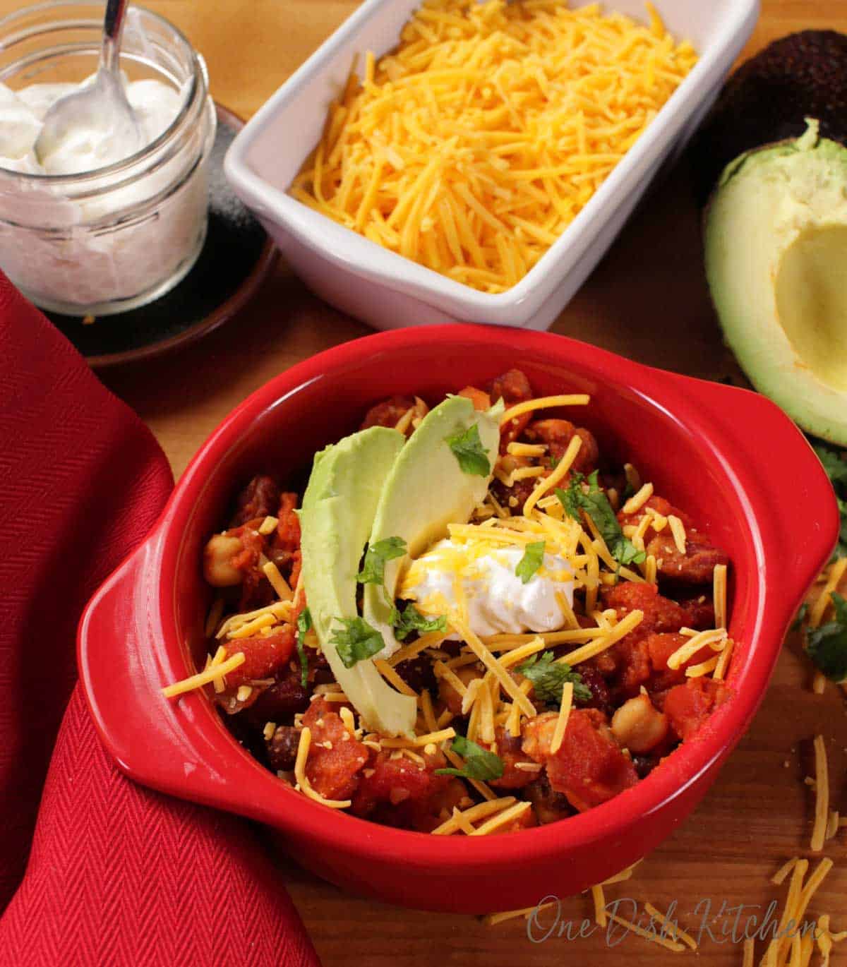 An overhead view of a circular baking dish of vegetarian chili topped with two slices of avocados, shredded cheese, and a dollop of sour cream next to a small jar of sour cream, shredded cheese, and half of an avocado. 