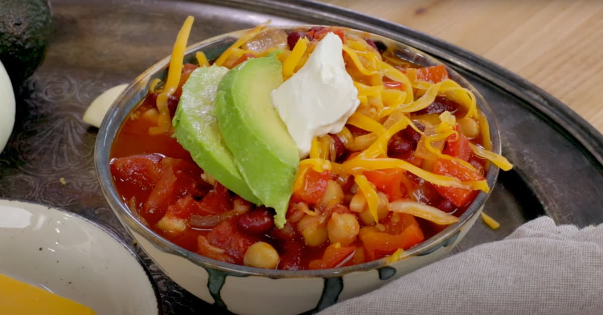 a bowl of vegetable chili topped with sour cream, cheese, and avocados on a silver tray