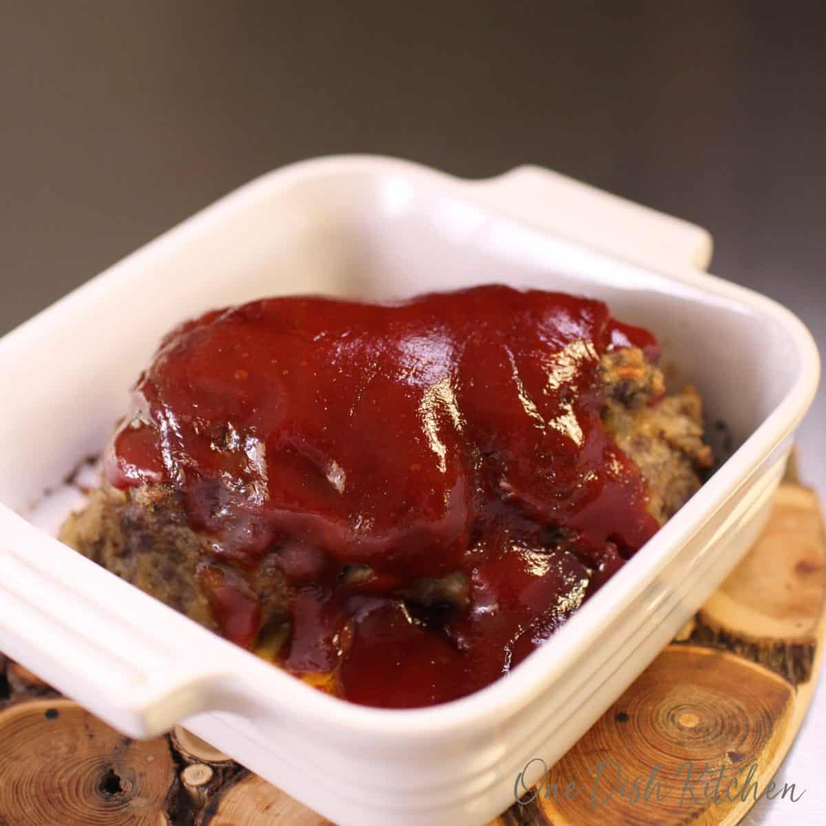 Meatloaf topped with sauce in a small baking dish on a wooden trivet.