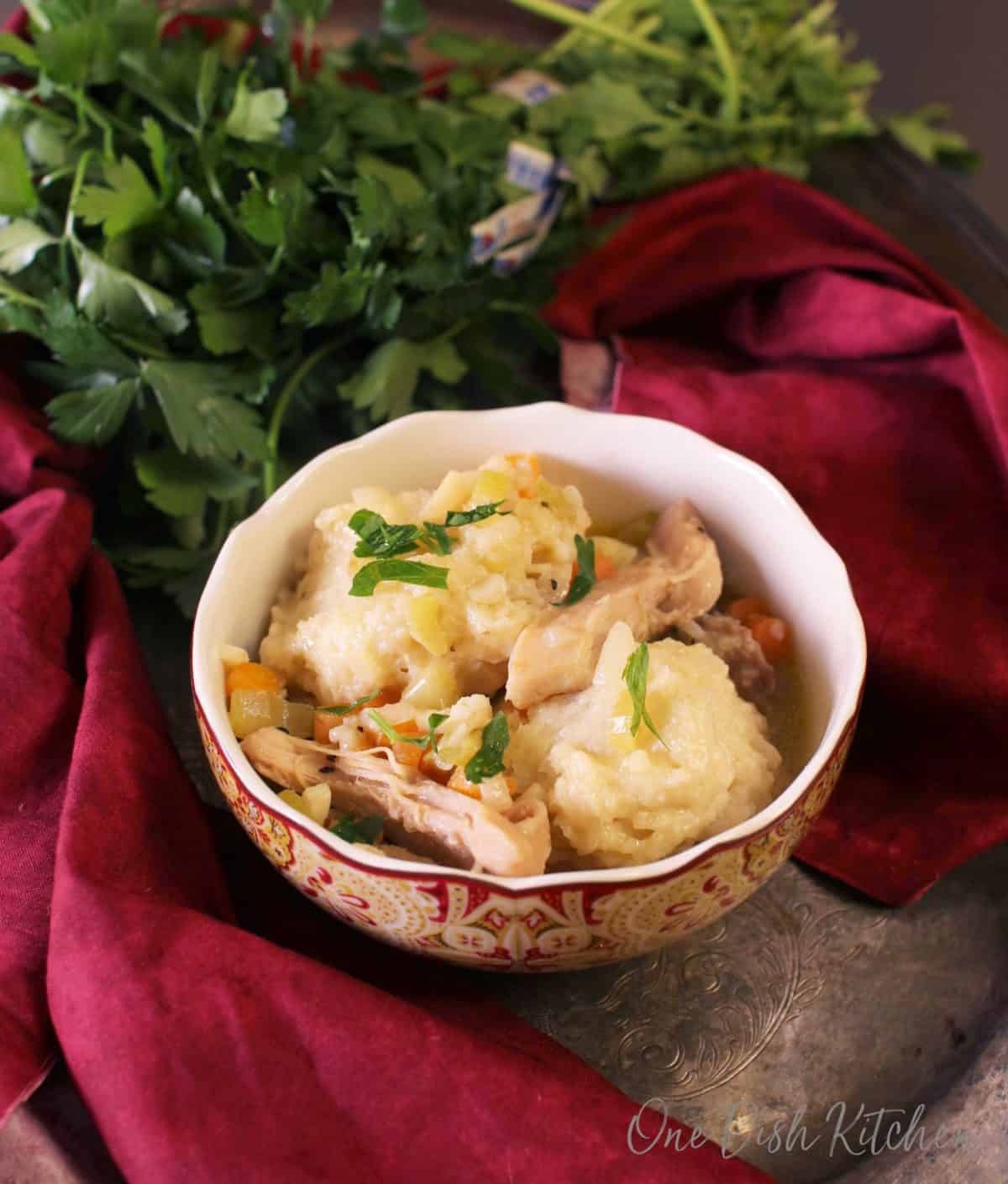 A bowl of chicken and dumplings on a metal tray with a bunch of parsley and a red cloth napkin 