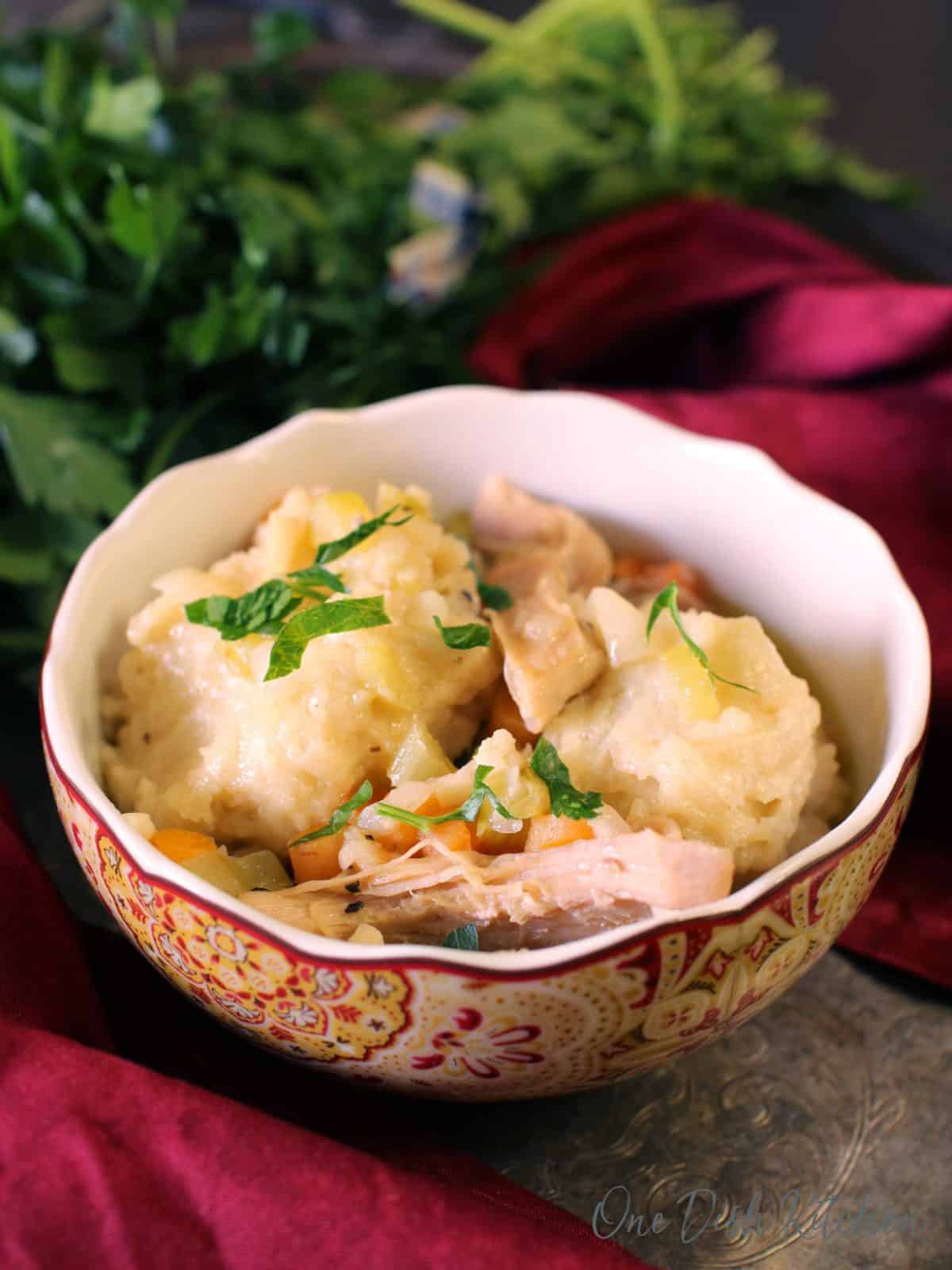 A bowl of chicken and dumplings on a metal tray next to a red cloth napkin and a bunch of parsley 