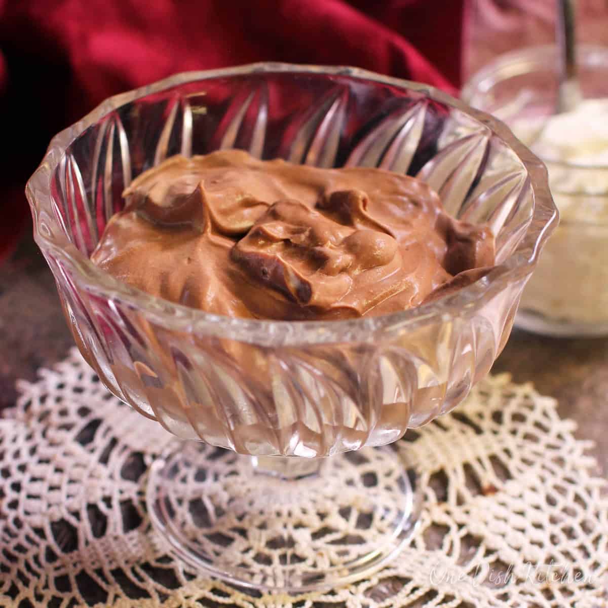 Chocolate mousse in a dessert glass next to a jar of whipped cream and a red cloth napkin all on a metal tray.