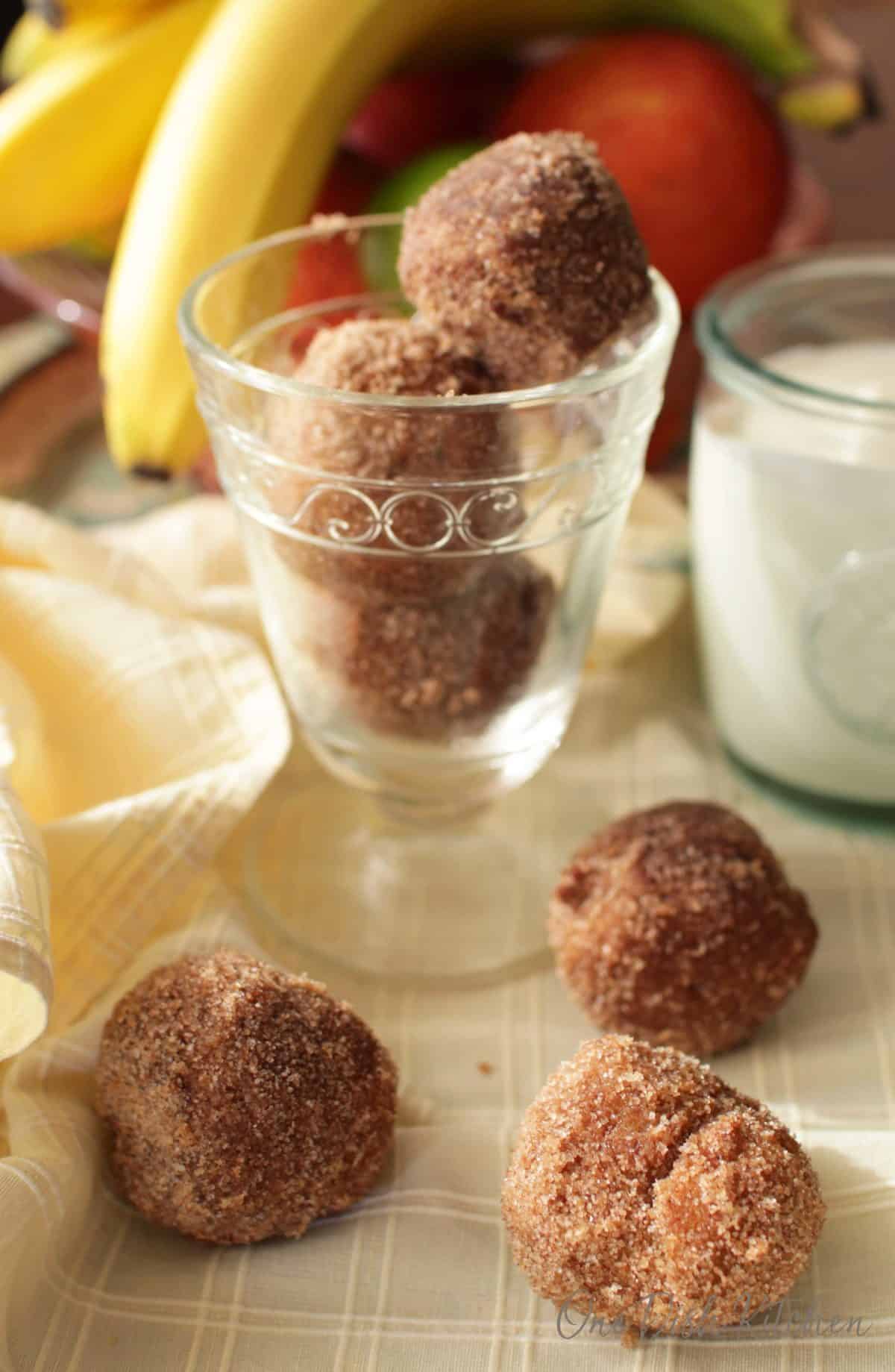 A dessert glass filled with three donut holes next to three donut holes scattered on a cloth napkin all next to a glass of milk