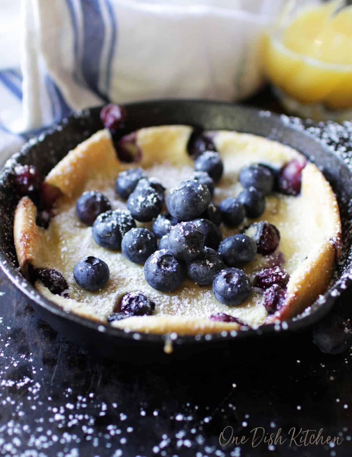 A dutch baby pancake topped with blueberries and powdered sugar next to a glass of orange juice.