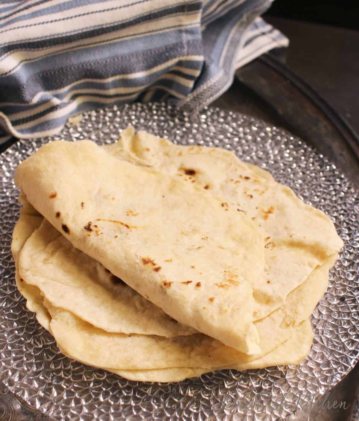 A stack of homemade flour tortillas on a plate placed on a metal tray with a blue and white striped cloth napkin.