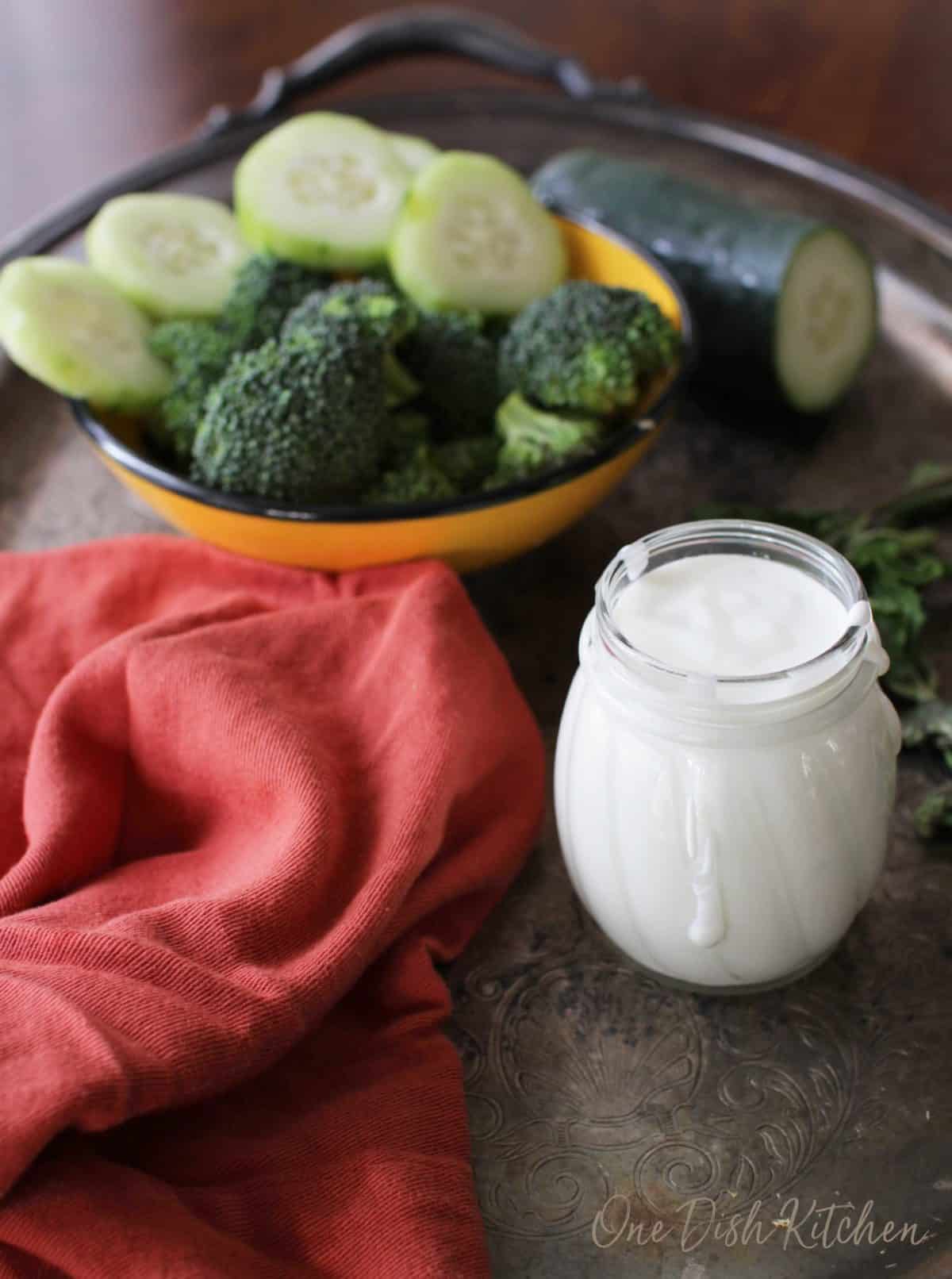 A small jar of goat cheese salad dressing on a metal tray with a red cloth napkin and a bowl of cucumber slices and broccoli florets 
