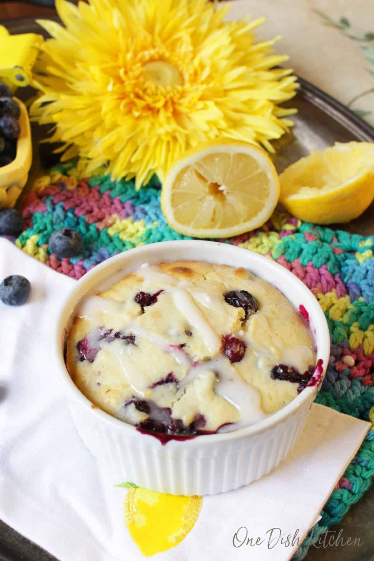 A lemon blueberry muffin in a ramekin topped with sugar glaze on a metal tray surrounded by blueberries, two lemon halves, and a big yellow flower.