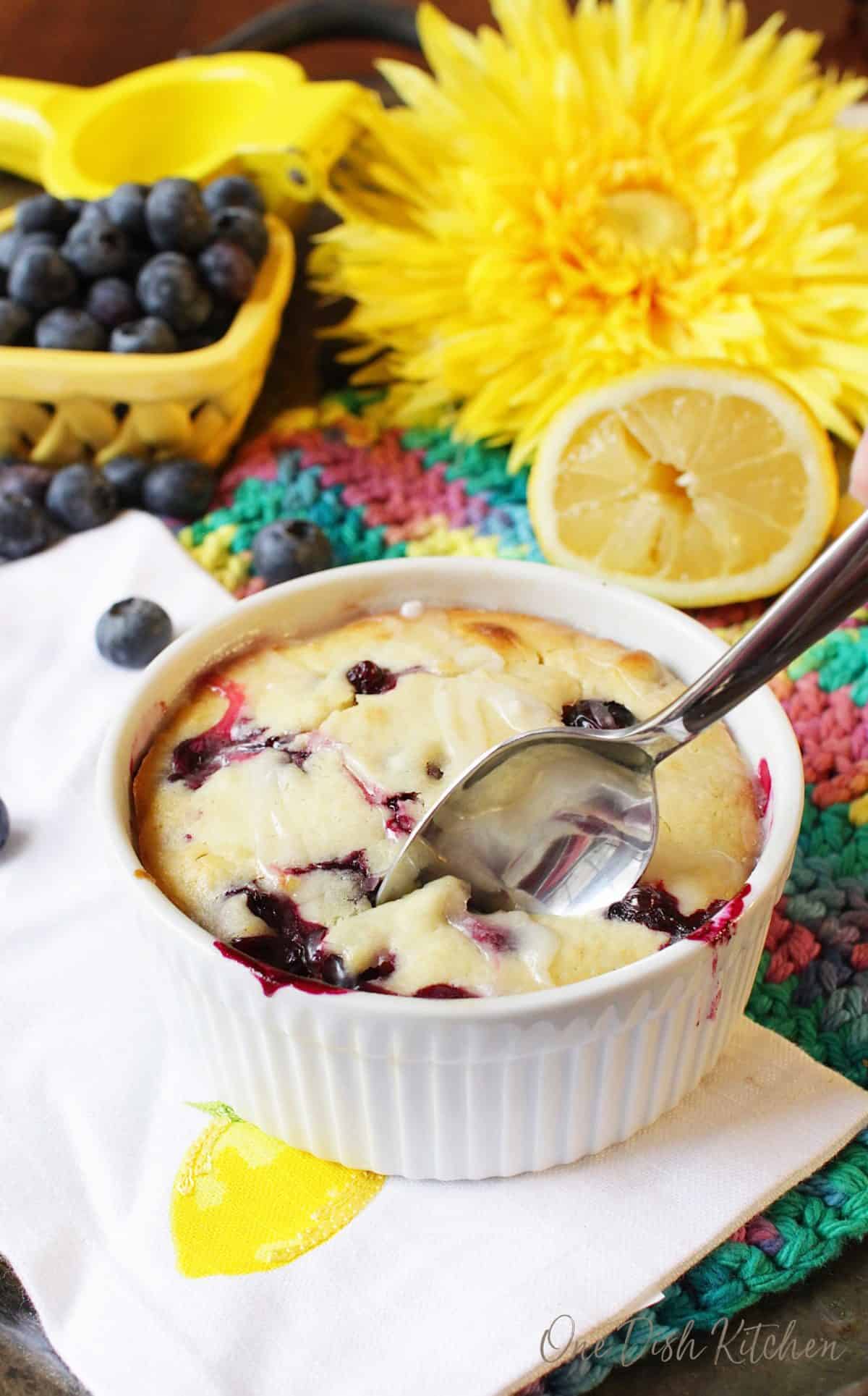 A spoonful of lemon blueberry muffin in a ramekin topped with sugar glaze on a metal tray surrounded by blueberries, half of a lemon, and a big yellow flower.