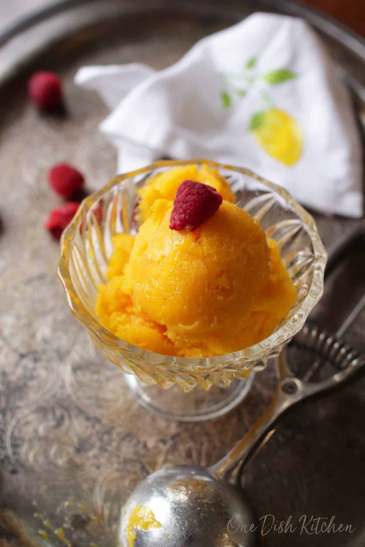 Mango sorbet topped with a raspberry in a dessert glass next to an ice cream scoop on a metal tray with scattered raspberries.