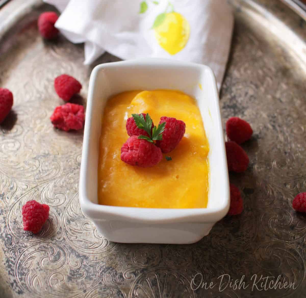 A small loaf pan filled with mango sorbet topped with raspberries on a metal tray with scattered raspberries.
