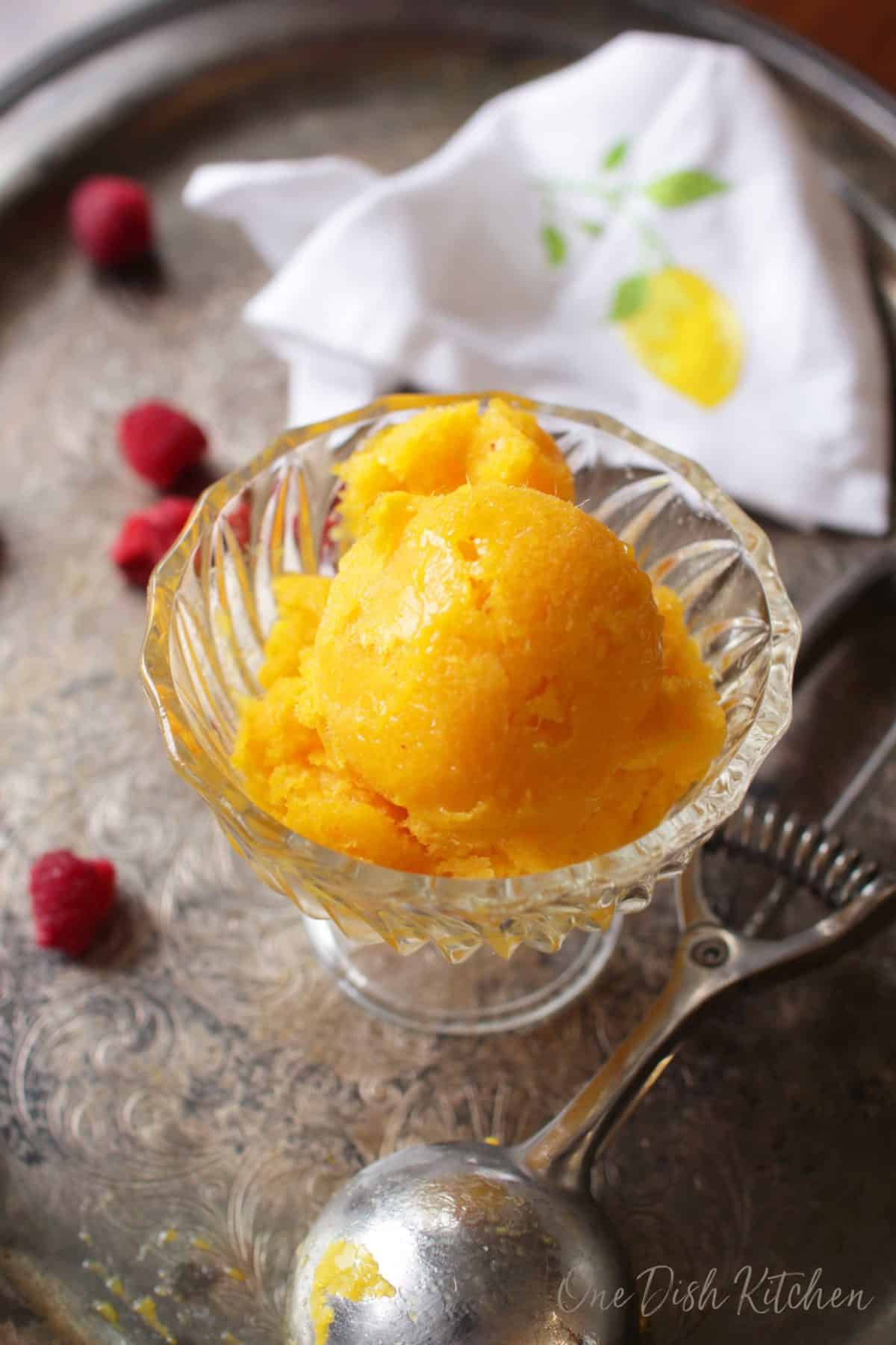 Mango sorbet in a dessert glass on a metal tray next to an ice cream scoop and scattered raspberries.