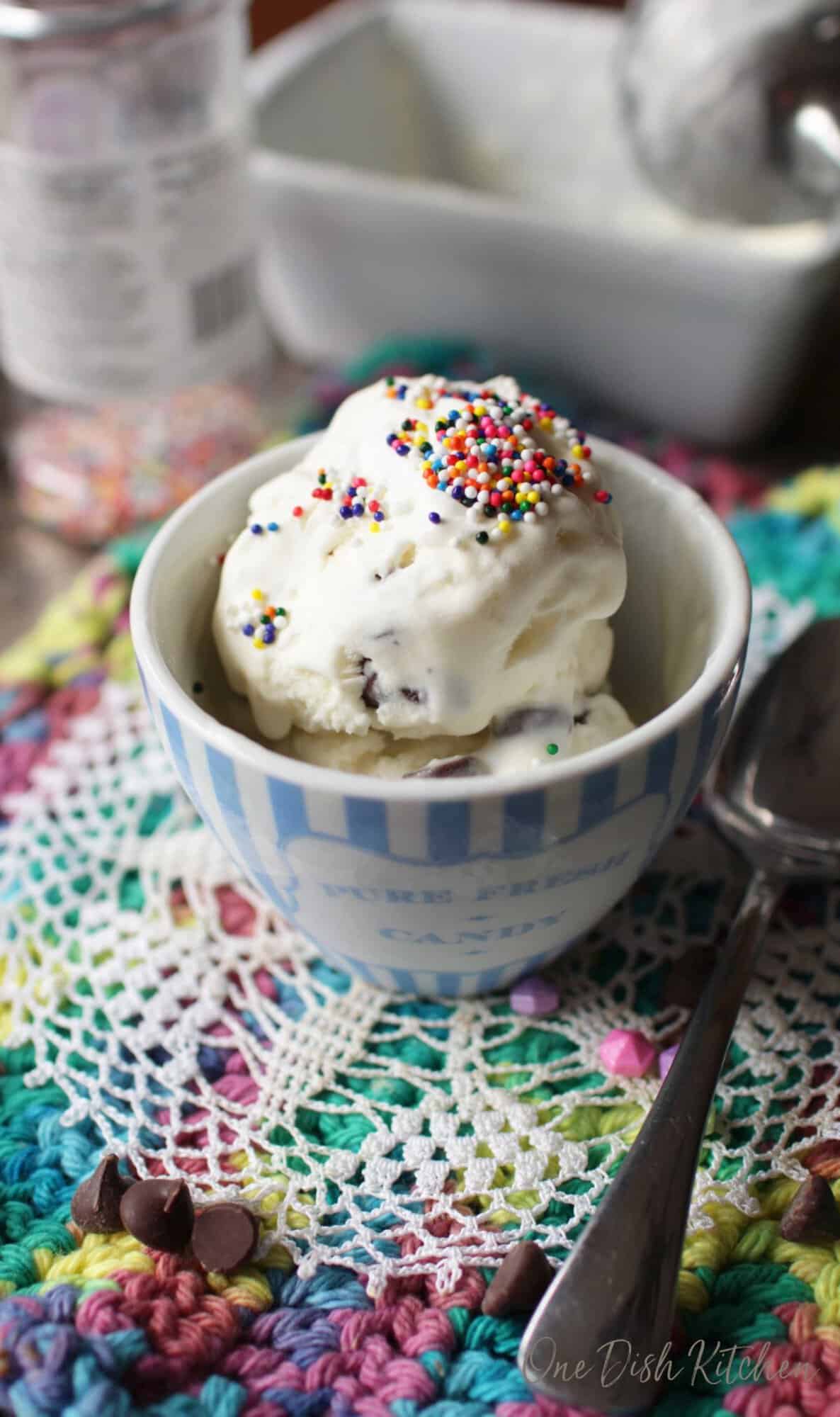 A small bowl of mint chocolate chip ice cream topped with rainbow sprinkles on a colorful surface next to a spoon and scattered chocolate chips