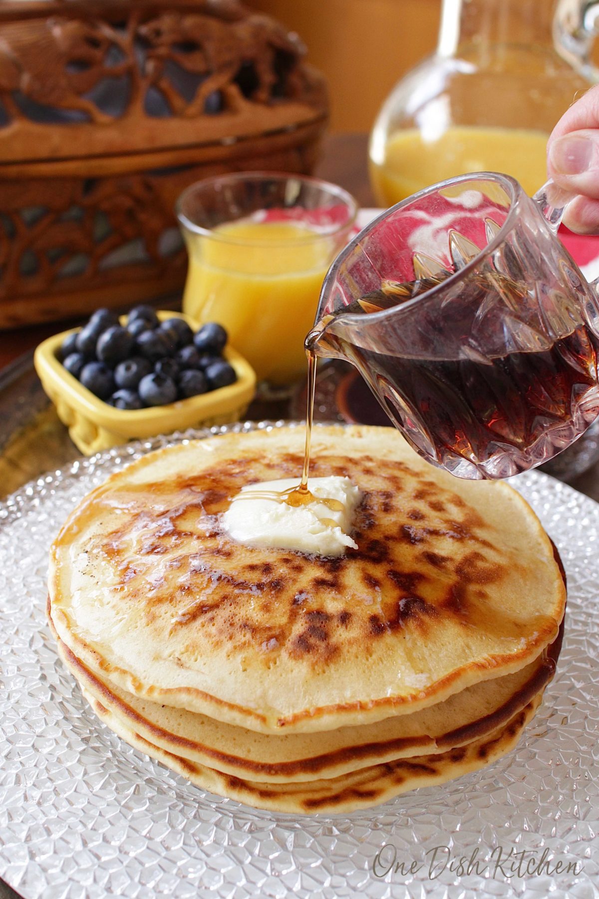 Pouring syrup over stack of pancakes on a plate topped with melting butter.