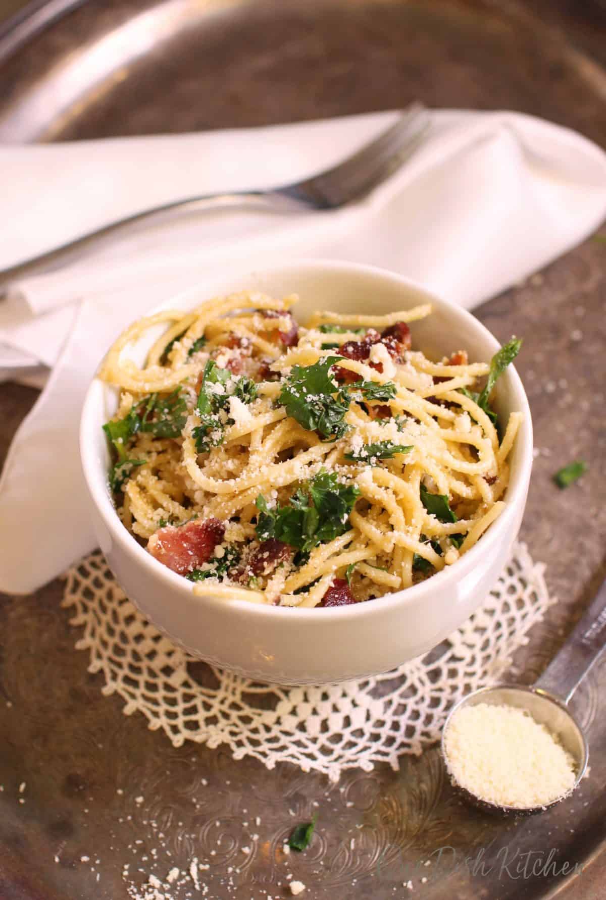 An overhead view of a bowl of pasta carbonara topped with chopped parsley and parmesan cheese next to a white cloth napkin, a fork, and measuring spoon of grated parmesan cheese all on a metal tray.