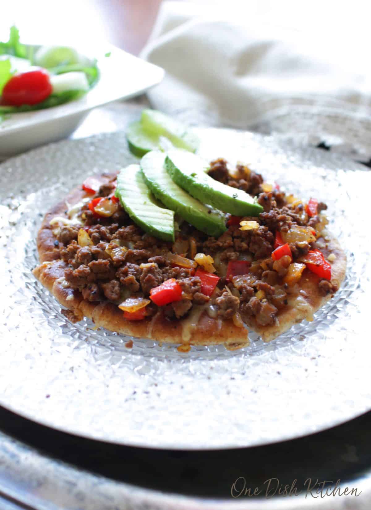 A pizza made on pita bread made with ground beef and topped red peppers, cheese, onions, and three avocado slices.