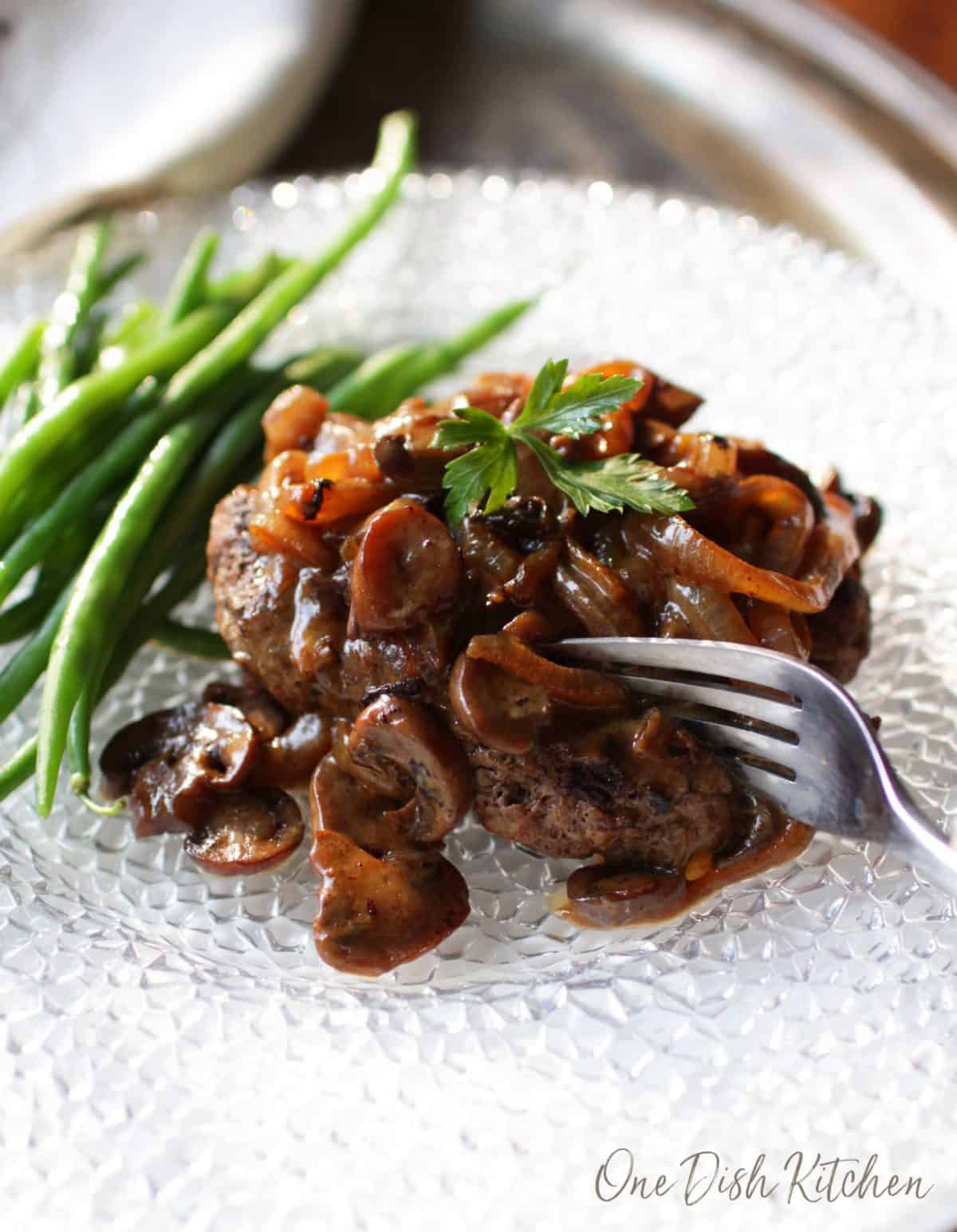 A fork going into a salisbury steak topped with mushroom and onion gravy with a side of green beans all on a large plate