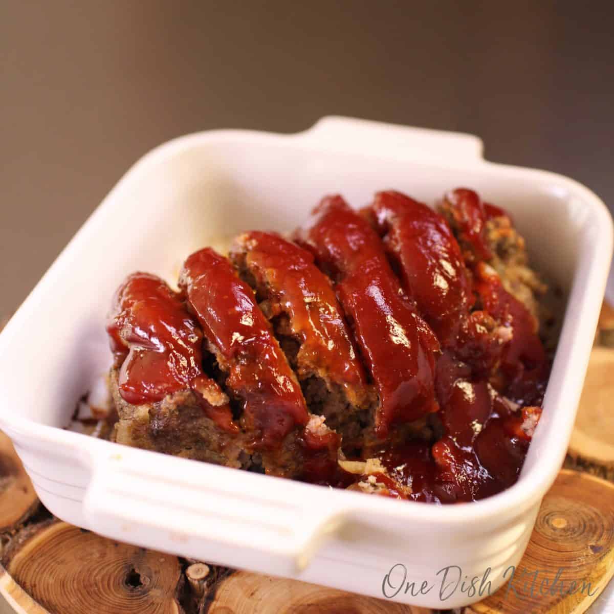 Slices of meatloaf topped with sauce in a small baking dish on a wooden trivet.
