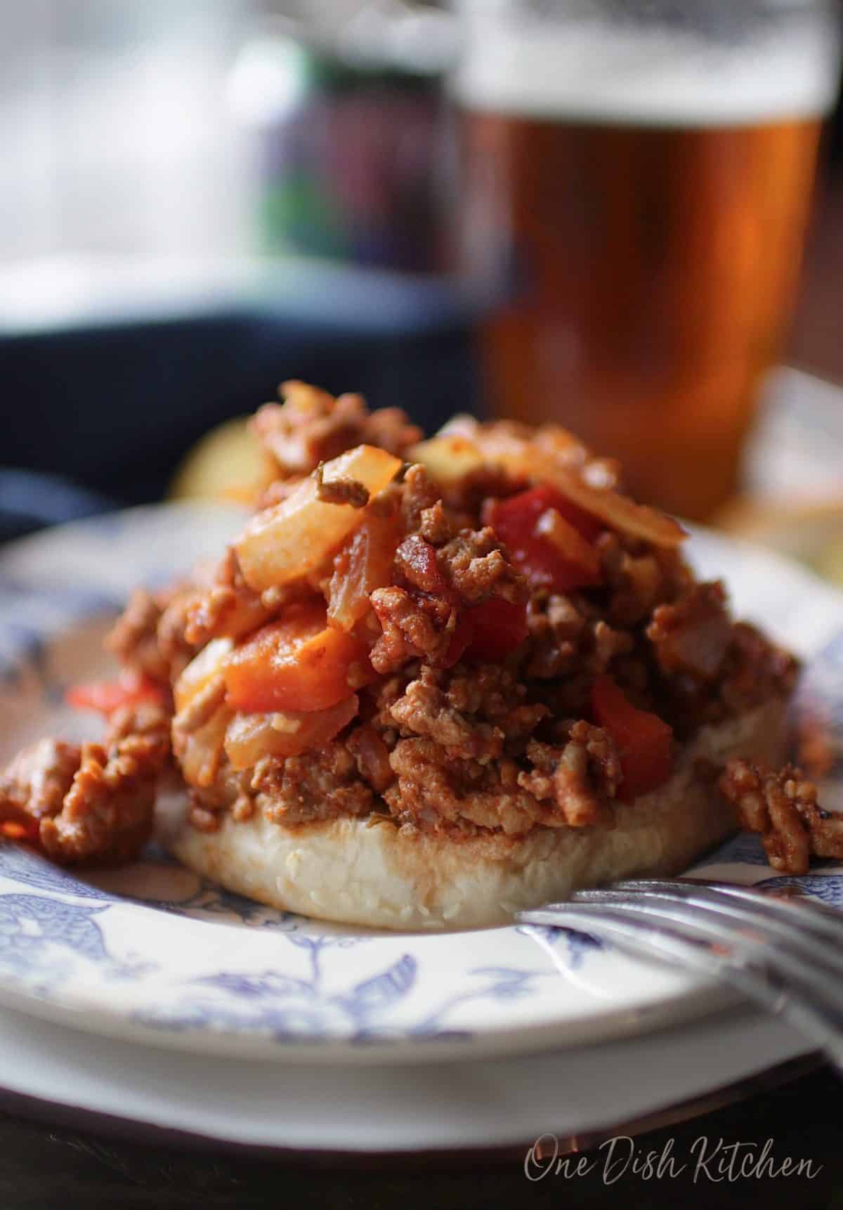 An open faced sloppy joe without the top bun on a plate next to a pint glass of beer.