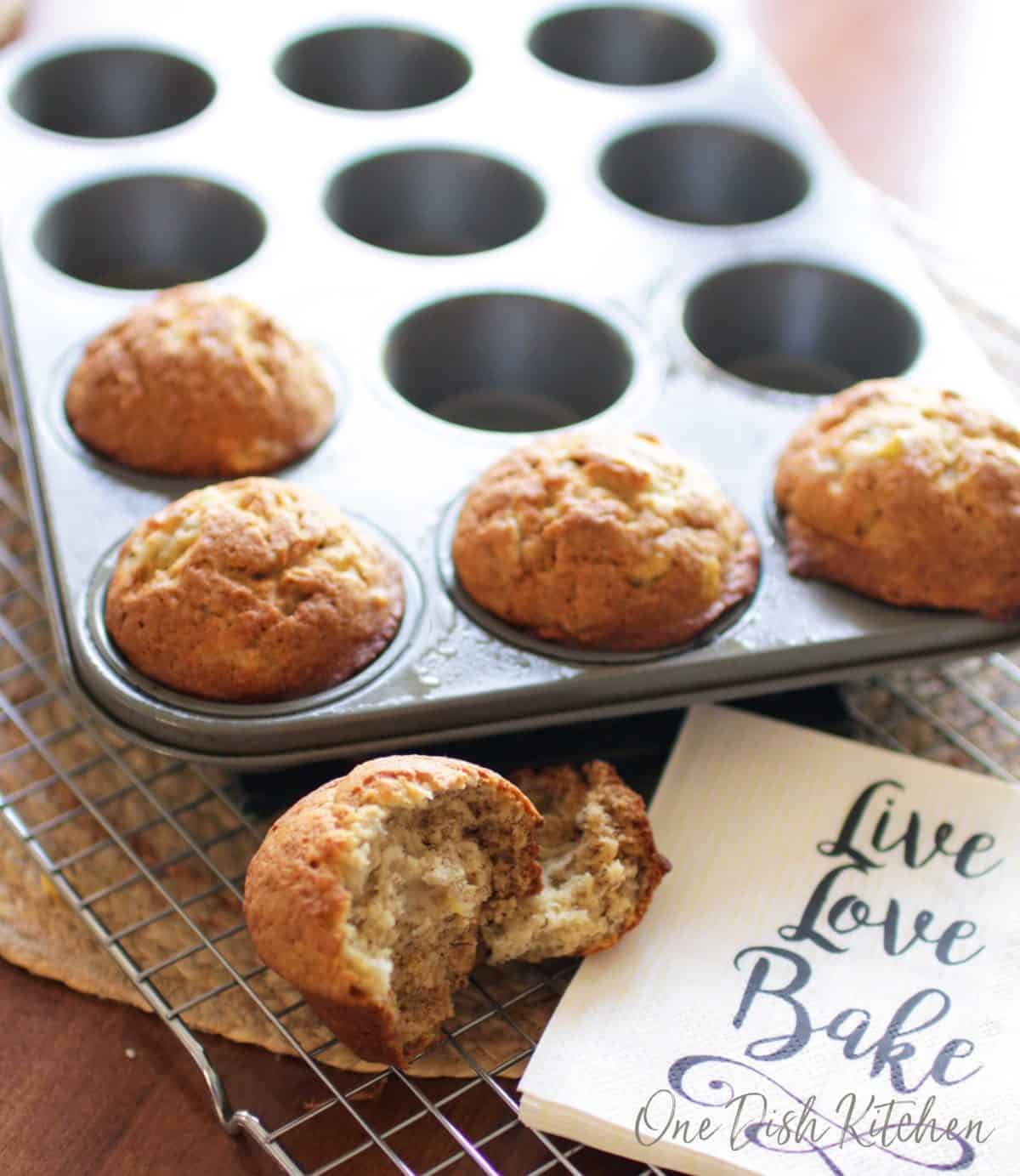 Four banana muffins in a muffin tin and a muffin split in half on a cooling rack.