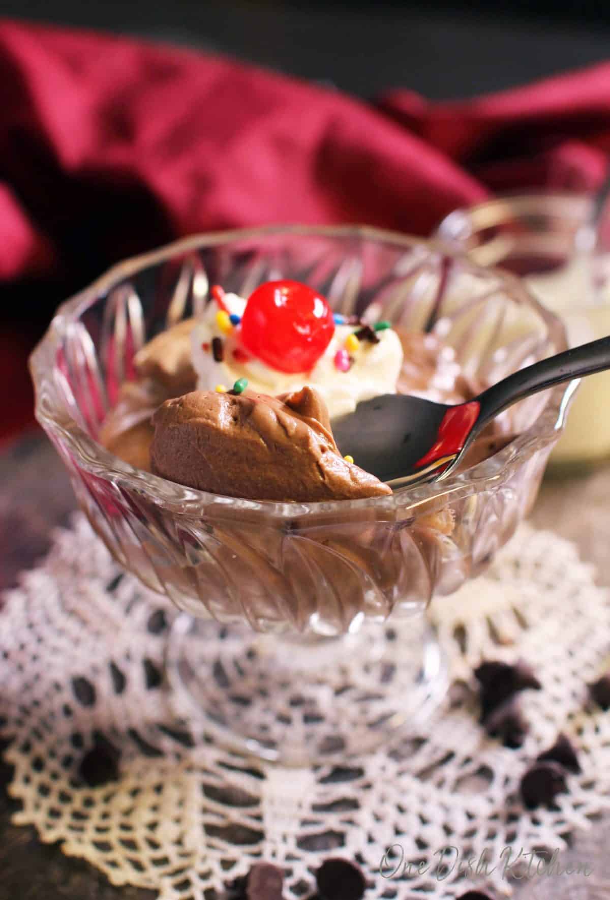 A spoonful of chocolate mousse being scooped out of a dessert glass topped with whipped cream, a cherry, and rainbow sprinkles on a metal tray with scattered chocolate chips, a small jar of whipped cream, and a red cloth napkin