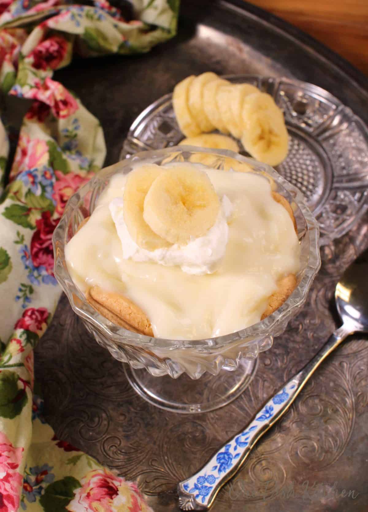 Banana cream pie in a dessert glass topped with whipped cream and sliced bananas on a metal tray with a floral cloth napkin, a small plate of sliced bananas, and a spoon