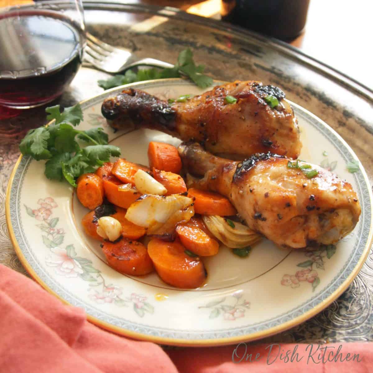 Two bbq chicken drumsticks on a plate with chopped carrots and garlic next to a glass of red wine all on a metal tray.
