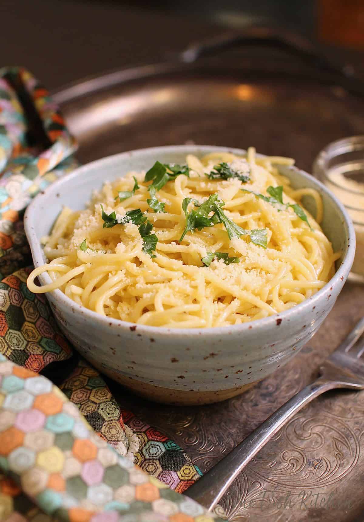 Spaghetti noodles topped with parmesan cheese and chopped parsley in a bowl next to a fork on a metal tray.