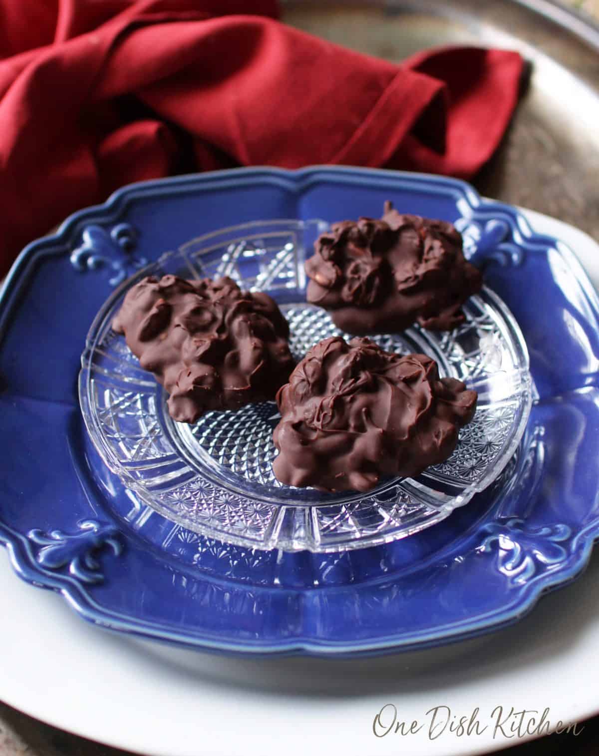 Three chocolate candies on a clear plate on top of a big blue plate on a tray with a red cloth napkin.