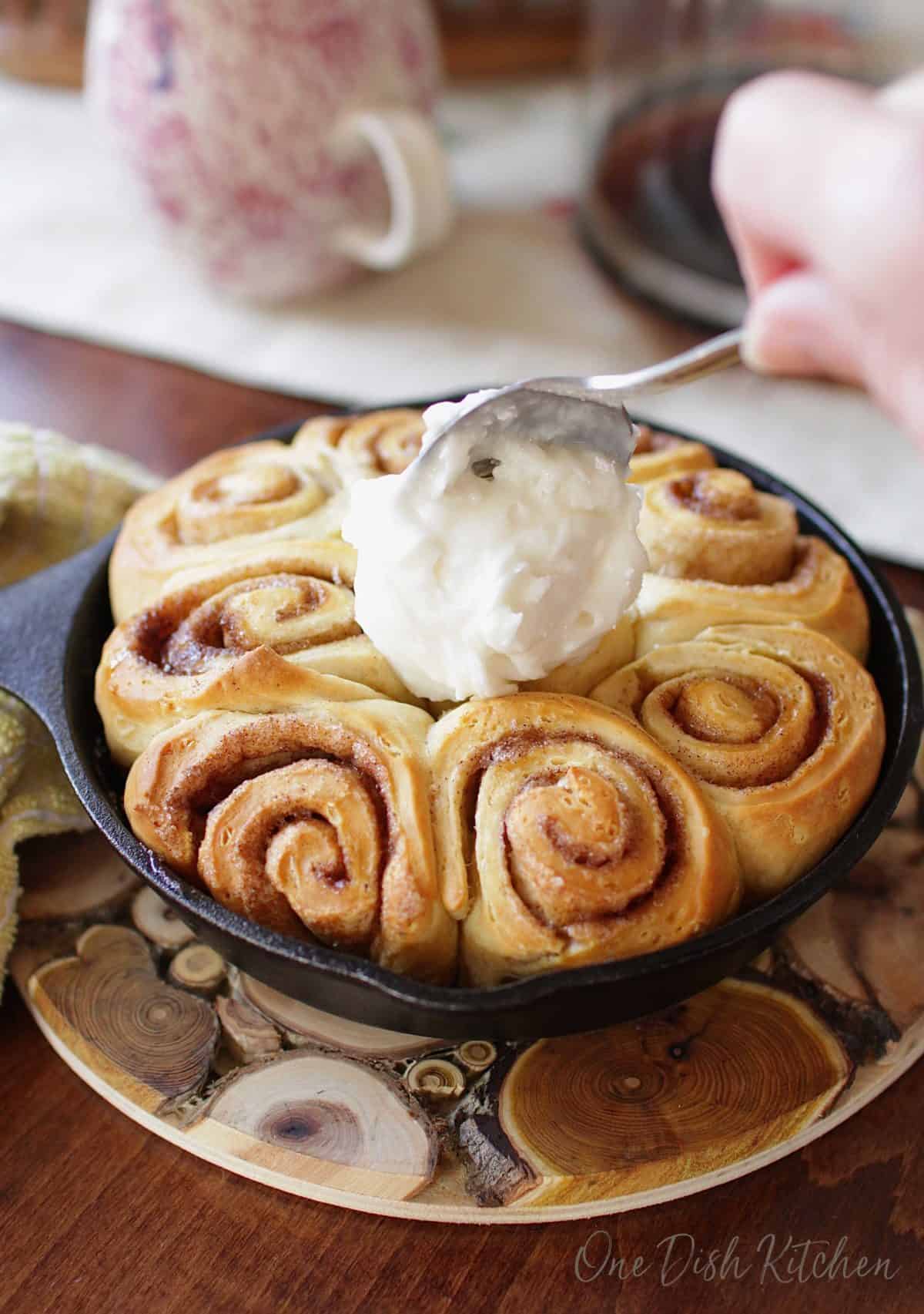 Spreading cream cheese frosting over homemade cinnamon rolls in a small skillet with the back of a spoon.