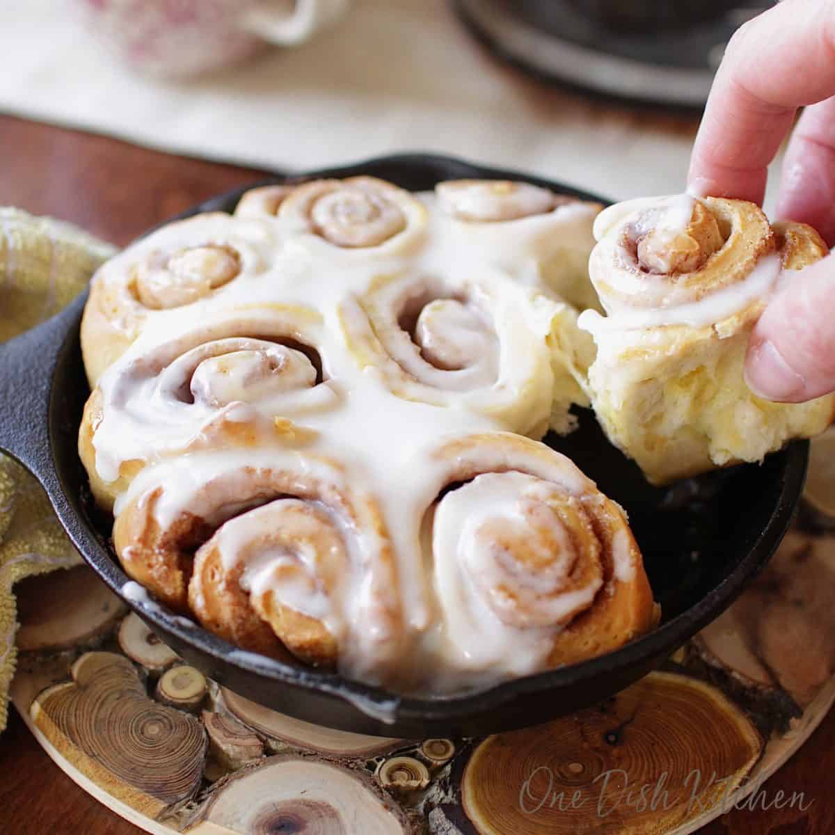 Pulling a small cinnamon roll out of a small skillet filled with cinnamon rolls topped with frosting on a trivet.