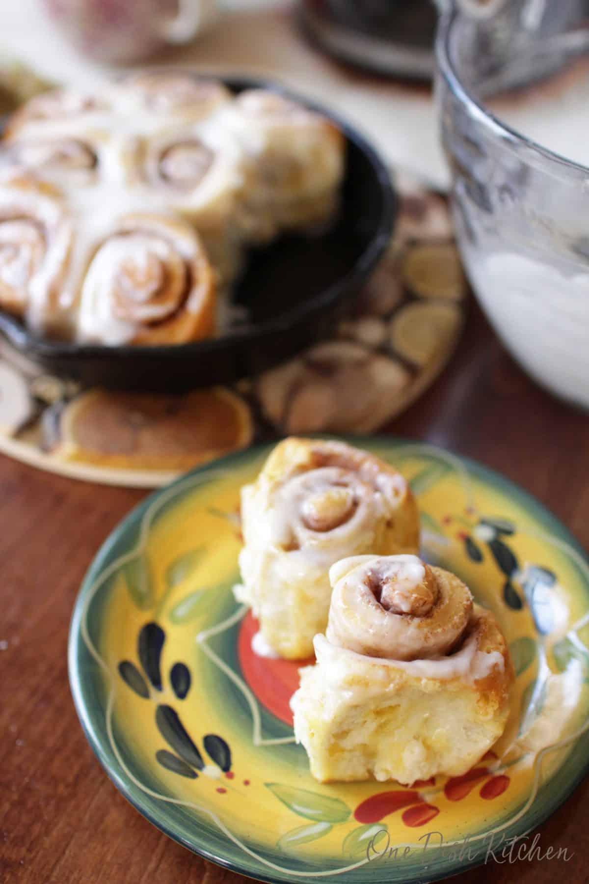 Two mini cinnamon rolls on a plate next to a small cast iron skillet filled with cinnamon rolls and a bowl of frosting.