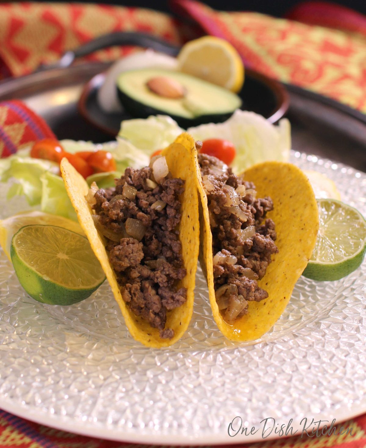 Two taco shells on a plate filled with ground beef and onions with lime halves on both sides of the tacos and a small salad on a metal tray with small plate of half of an avocado and half of a lemon in the background.