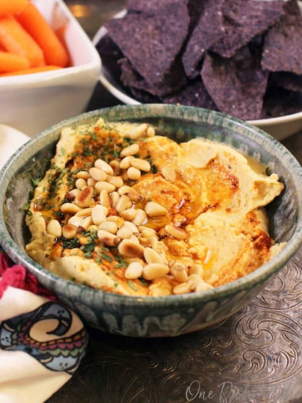 A bowl of hummus topped with toasted pine nuts, olive oil, and paprika next to a bowl of blue corn tortilla chips and a small bowl of baby carrots all on a metal tray.