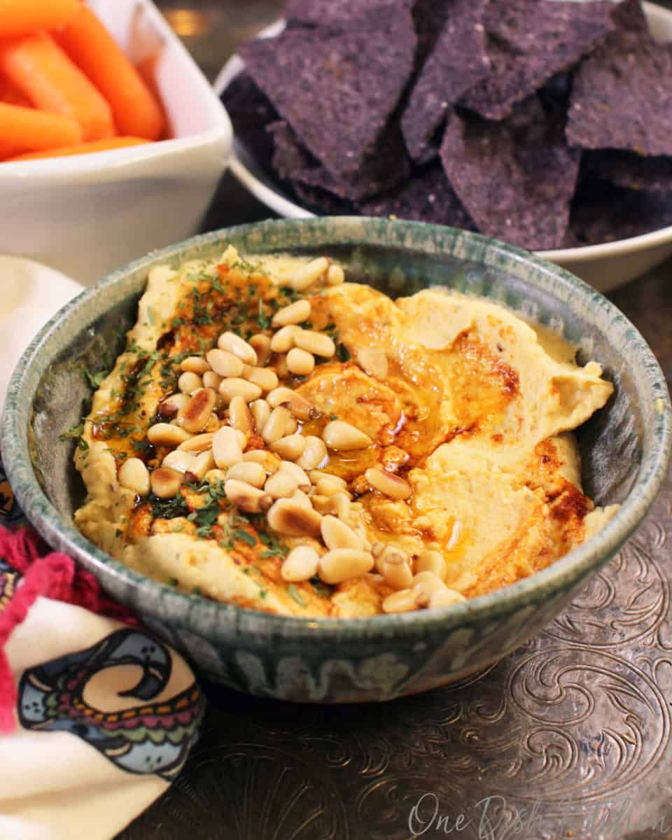 A bowl of hummus topped with toasted pine nuts, olive oil, and paprika next to a bowl of blue corn tortilla chips and a small bowl of baby carrots all on a metal tray.