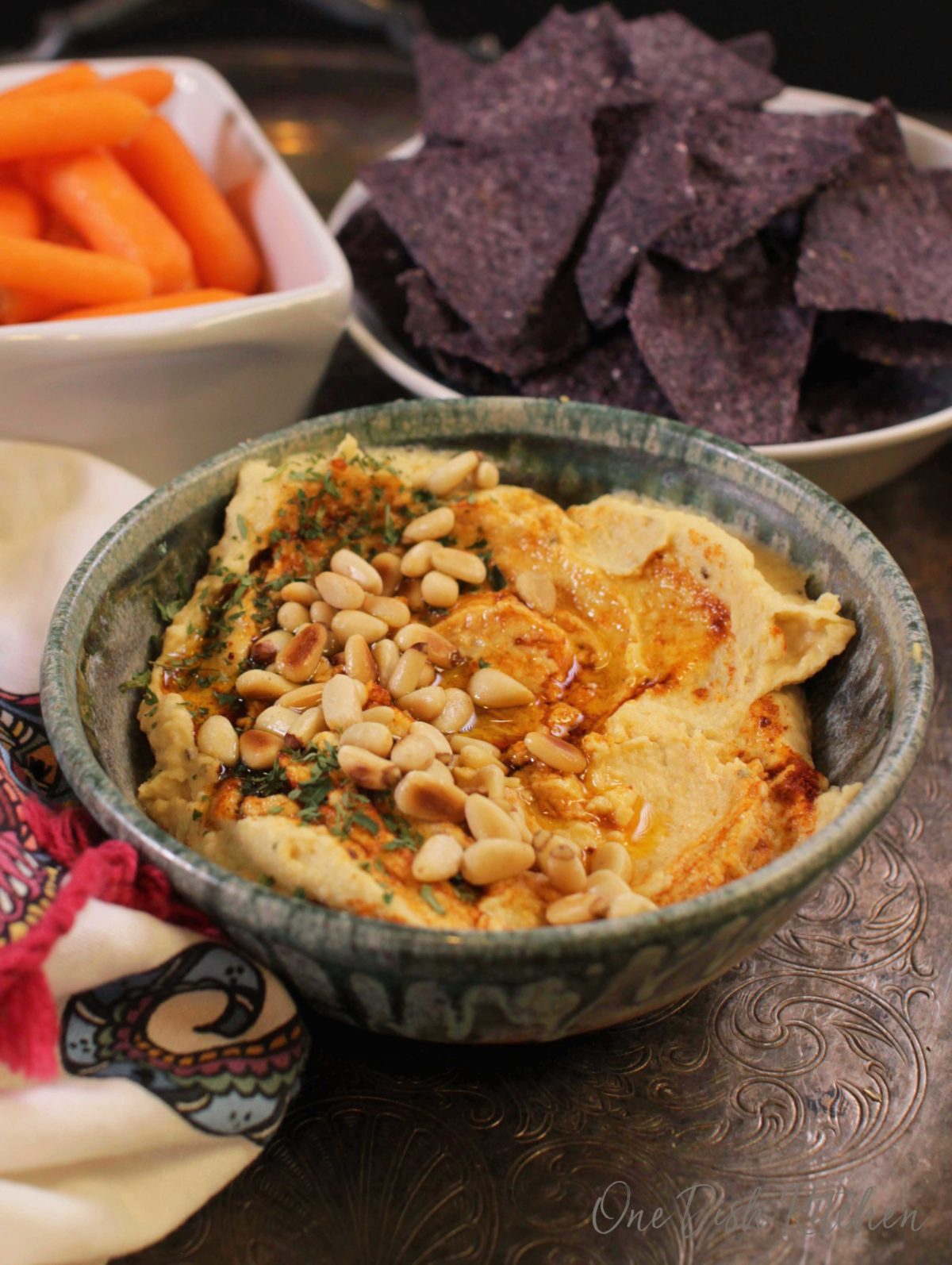 A bowl of hummus topped with toasted pine nuts, olive oil, and paprika next to a bowl of blue corn tortilla chips and a small bowl of baby carrots all on a metal tray.