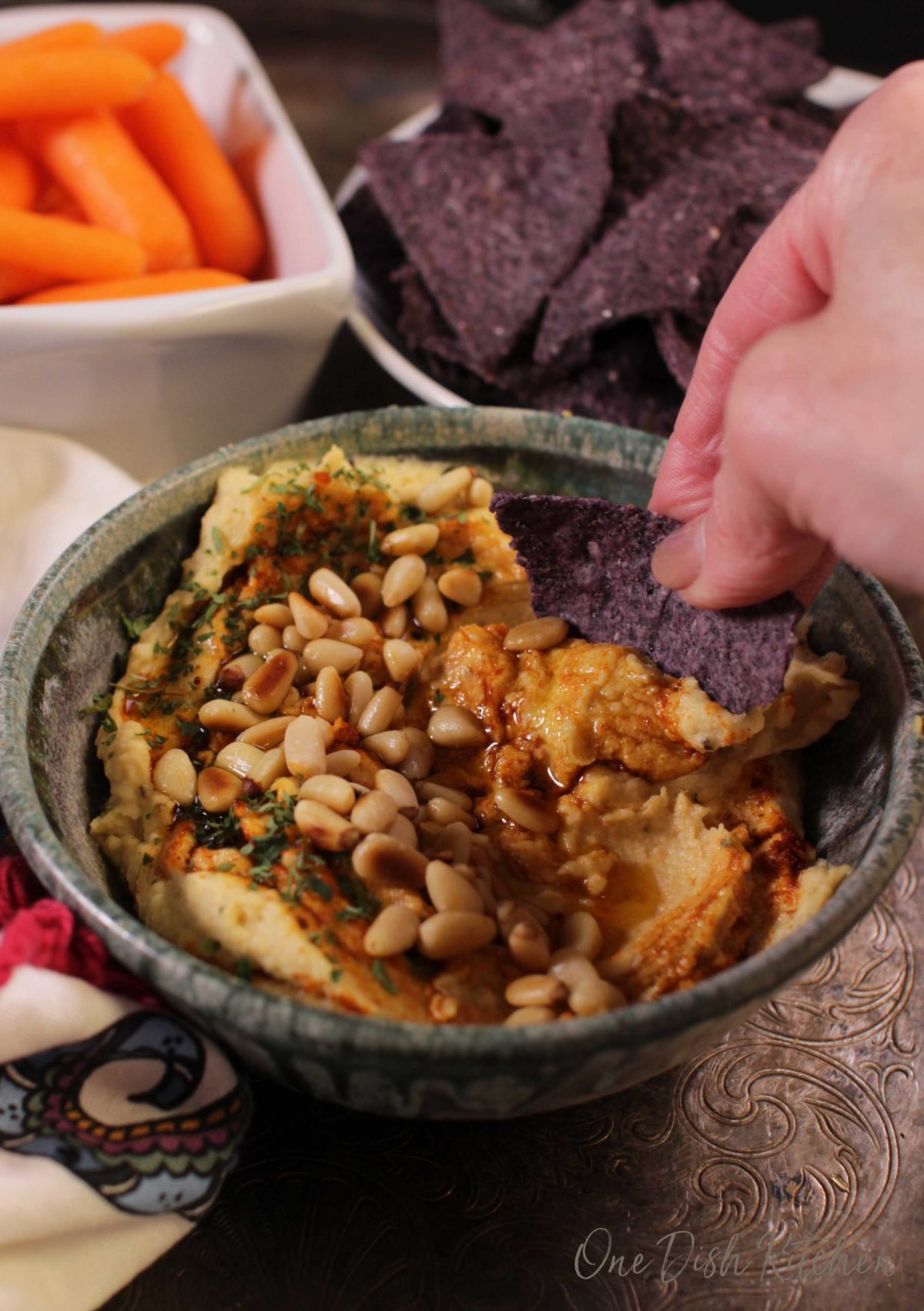 A blue corn chip scooping out hummus from a bowl that is topped with toasted pine nuts, olive oil, and paprika next to a bowl of blue corn tortilla chips and a small bowl of baby carrots.