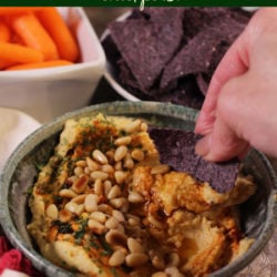 a blue corn chip dipped into a bowl of hummus topped with toasted pine nuts and olive oil.